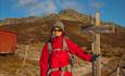 Hiker in a red jacket outside the cabin Storeskag at the sign pointing the way towards the summit of Skaget.