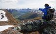 Eine Frau mit Rucksack sitzt auf einem Felsen und genießt eine gandiose Aussicht in ein tiefes Tal mit einem See im Talgrund.