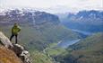 A man stands on a rocky outcrop in a steep hillside and enjoyes a magnificent view into a steep valley with a lake on the bottom.