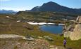 A little girl on a path in the mountains with a small waterbody along the trail and a massive mountain in the background.