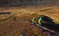 Children run along a path between low heather vegetation in the mountains in autumn. In the background the valley bottom with a mountain farm can be s