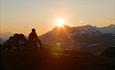 A man sits on a rock and looks into the sunset behind a high mountain.