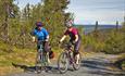 Two cyclists are struggling up the last bit of a steep climb on a gravel road. Snow covered mountains can be seen on the horizon.