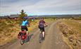 Two cyclists cycle on a farm road on a mountain plateau past farm buildings, enjoying a far view.