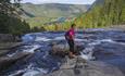 A woman stands on a naked rock in a river before it drops into a waterfall into the valley seen beyond.