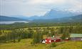 Rød stølshytte på langestølen med grønne lier. I bakgrunnen ses en flik av vannet Storfjorden og det mektige fjellet Synshorn.