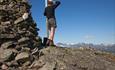 A woman stands next to the summit cairn and looks out to distant mountains.