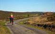 Syklist på stølsvei gjennom en stølsgrend med røde hus og snødekte fjell i horisonten.