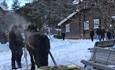 A horse with a sleigh on a farm yard in the snow with persons standing around.