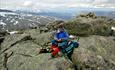 A man sits at the summit of Rasletinden on a dry, but overcast day. In the far background, Sjodalen Valley and other 2K peaks are visible.