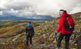 Two hikers on their way down from Rundemellen through rocky terrain. Green hills and low clouds in the background.