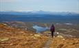 A person hikes on a path towards a stunning view with lakes, several hilly ridges and snow-capped high mountains in the rear.