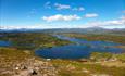 Utsikt over innsjø-landskap med knauser og holmer, noe granskog og fjell i himmelrand.