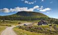 Farm road with parking space, a house and spruce forest in front of a mountain.