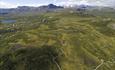 Aerial image of a mountain plateau with a gravel trail for terrain cyclists leading around a swamp. High mountains in the background.