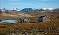 Tomashelleren en flott høstdag. To gråfargede hytter ved et lite vann, og ruvende fjelltopper i bakgrunnen.