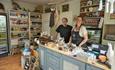 The owners of the store standing behind the counter of the small, cosy shop.The shelves are filled with delicious local food.