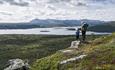 Kvinne og barn står på liten stein og skuer utover et storslått landskap, med innsjø og fjell i bakgrunnen.