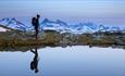 Crystal clear lake, woman walking by the lake is reflected by the water surface. High, pointed mountains in the far background. Purple morning light b