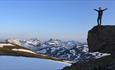 Clear sky, hiker standing on top a rock at the right while enjoying wild mountains with snow spots in the background.