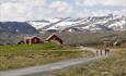 Fire syklister på en grusvei i fjellet, med røde stølshus og snødekt høyfjell bakenfor.