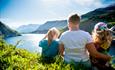 A family sits in a hillside and enjoys the view over a lake surrounded by mountains.