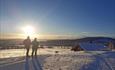 Zwei Personen in einer Skiloipe bei einer Hütte im Gegenlicht der untergehenden Sonne