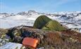 A green mountain tent on a bare patch in otherwise snow covered landscape with mountains in the background.