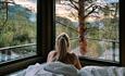 a woman lying in bed and looking out of the window into the forest