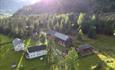 Dronenfoto des Hofes mit Gebäuden, großen Rasenflächen und angrenzendem Birkenwald. Sonnenstrahlen im gegenlicht erzeugen ein Mosaik aus Licht und Schatten.