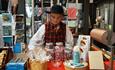 Ein junger Mann in Volkstracht hinter dem Ladentisch in einem Museumsladen mit Bonbons im Glas, traditionellen Strickwaren, Kunstkarten und Büchern.