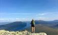 Wanderer auf einem Aussichtspunkt mit Ausblick über einen langgestreckten See umgeben von Bergen.