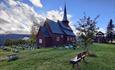 Stave church on a green lawn with a small rowan berry tree against the sun.