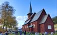 A red-brown stave church surrounded by a church yard with birch trees that start to get autumn-coloured leaves.