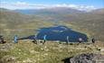 Fjellvandrere i rad og rekke med vann som ligger i dalen i bakgrunnen.