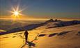 A mountain skier on a borad snowclad ridge with pointed peaks and the sun in the background.