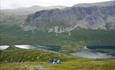 Four persons sit in the grass and enjoy a wild landscape with lakes and steep mountain sides.