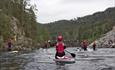 A group of stand-up boarders float down a river and underneath a bridge.