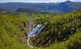 Doppelter Wasserfall in Schlucht mit grüner Vegetation. Grüne Wiesen und Berg im Hintergrund.