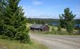 Open summer farm country at the tree line with a gravel road, a farm building and view over a lake with forest on the far side.