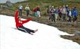 Ein Mädchen rutscht auf dem Po ein Schneefeld hinab, während der Rest der Wandergruppe zuschaut.