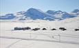 En liten fjellstue og et knippe hytter i et nedsnødd høyfjellslandskap fra fugleperspektivet. Skispor og skigåere utenfor hytta.