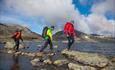 Tre vandrere steingår en elv i fjellet