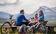 A family on a bicycle trip takes a break at a rest area with picknick table. Mountains in the background.