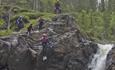 A group of persons with wetsuits and helmets jumps down a rock next to a waterfall in a river.