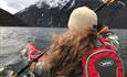 Girl in a red kayak og a fjord with black steep rock faces covered by a thin snow layer in the background.