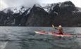 Girl in a red kayak og a fjord with black steep rock faces covered by a thin snow layer in the background.