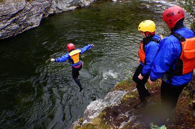 En person hopper fra en klippe ned i elva mens to andre ser på.