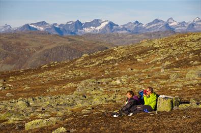 En familie tar en pause i gresset på et fjellplatå mellom mange lavdekte steiner på en solrik høstdag. Høye, spisse fjell i bakgrunnen.