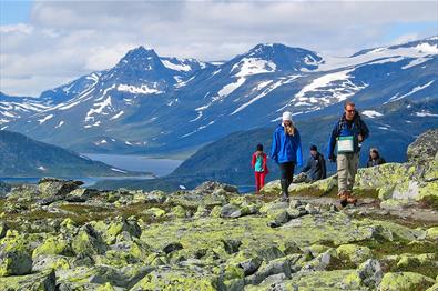 Hikers in the mountain pass a section with a lot of lichen-covered rocks. A lake and high mountains in the background.
